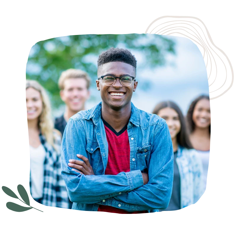 Smiling young teen in glasses standing confidently with arms crossed, with a group of people blurred in the background outdoors. life and leadership